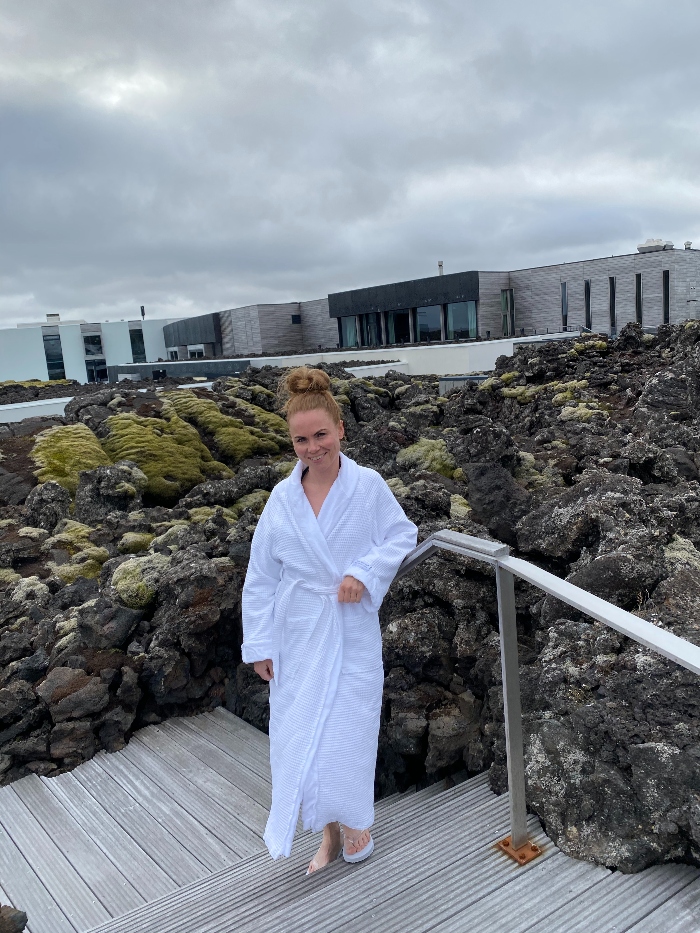 A woman visiting the Blue Lagoon posing in a bathrobe standing on a wooden stairs with lava field in the background
