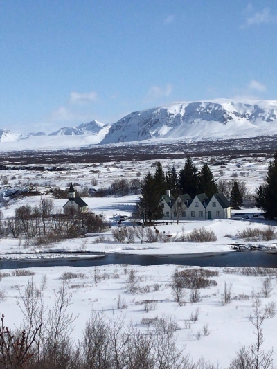 Snow covered ground with mountains in the far and a small river and old house in the foreground