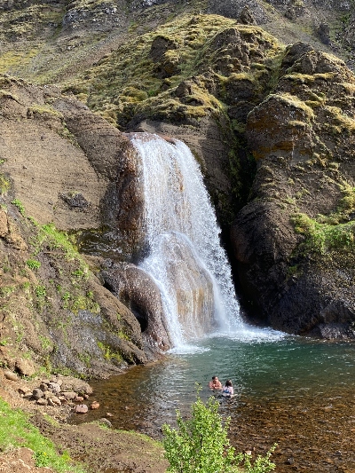 A waterfall falling from brown rocks with a little green on it and two people bathing in the water at the bottom