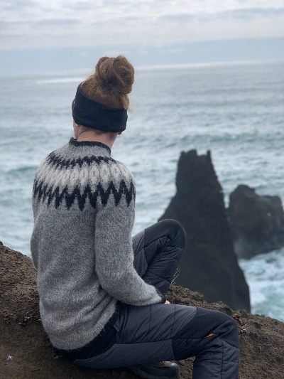 A woman wearing an icelander sitting on an edge of a cliff and watching over the sea 
