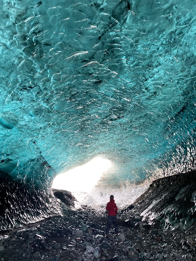 A woman in a red jacket standing in a middle of a huge and amazing blue ice cave