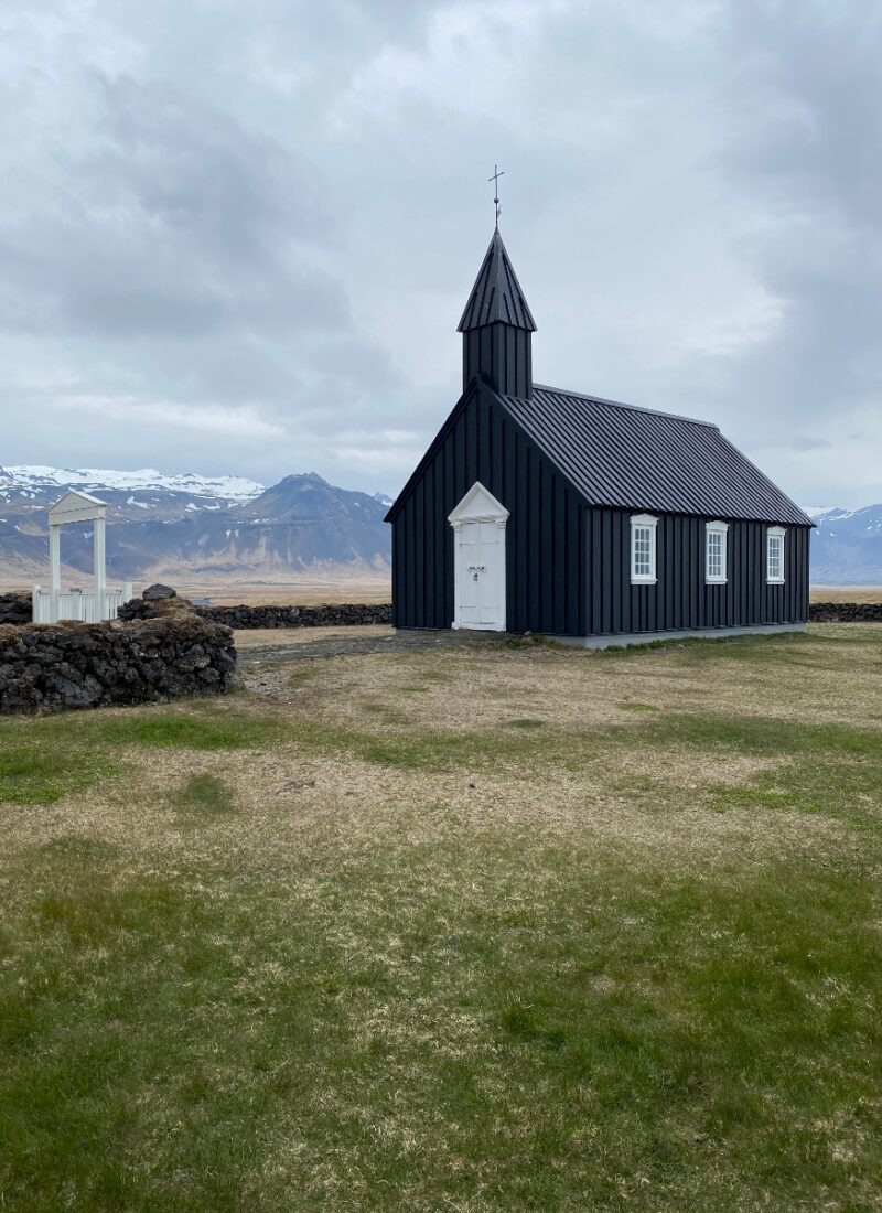 A black church made of timber standing by itself on a green grass with mountains in the background