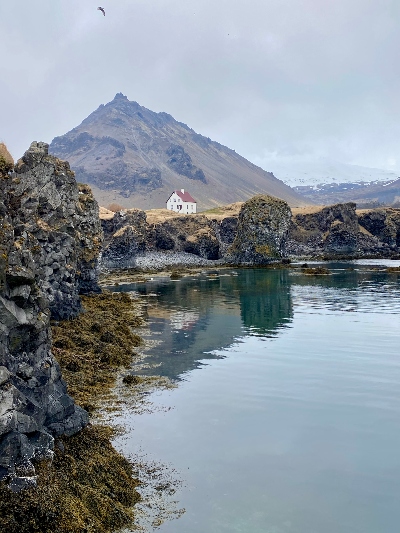 A small white house with a red roof standing alone by the sea surrounded by beautiful lava formations and with a stunning mountain in the background