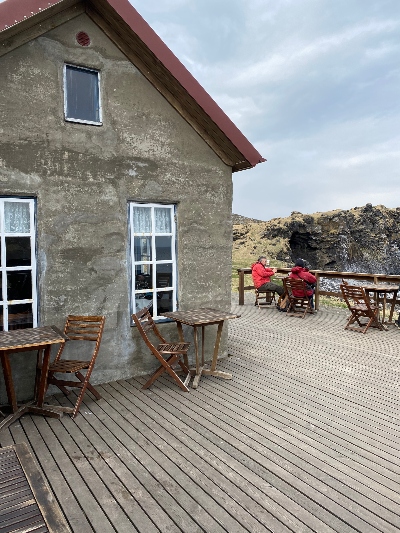 People in red jackets sitting by a table outside on the wooden patio at Fjöruhúsið Café at Hellnar