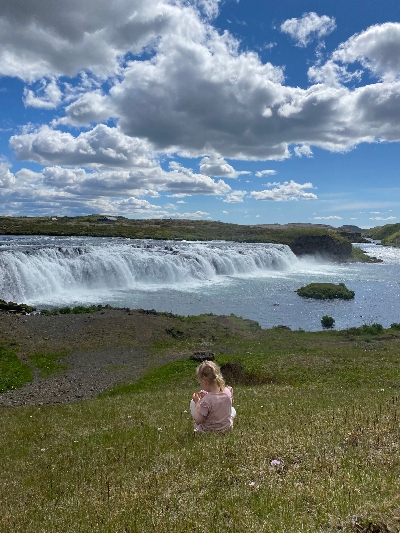 A little girl in pink sitting on the grass picking some flowers with a white waterfall in the background