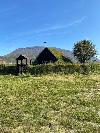 A small old turf church in Iceland surrounded with a circular turf wall with some mountains in the background on a sunny day