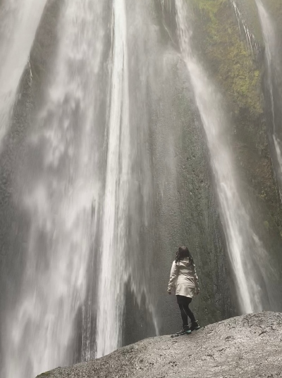 A woman in white rain jacket standing very close to a waterfall located in a cave