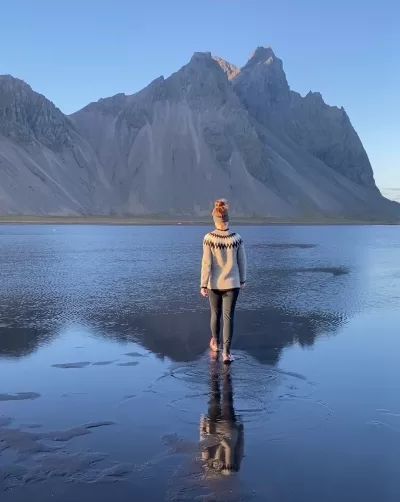 A woman walking on the reflection of Vestrahorn Mountain in the water in front of it