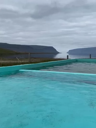 The turquoise Reykjalaug Pool in Iceland in the foreground and the ocean and some mountains in the background.
