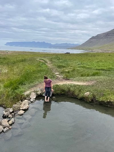 A woman dipping her toes into Reykjalaug Hot spring in Iclena with the ocean as a backdrop.